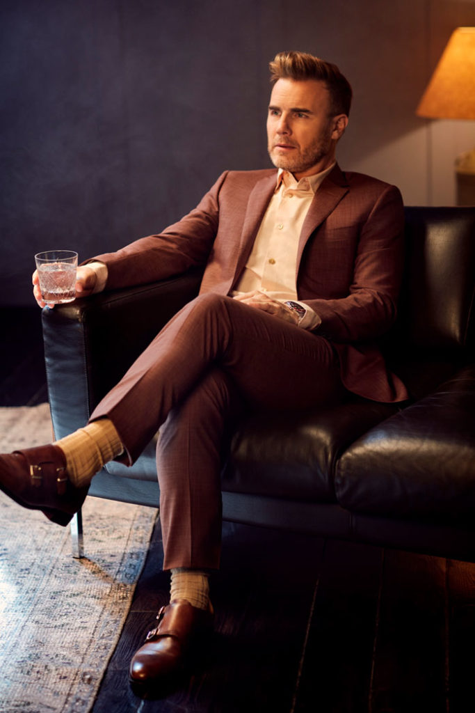 Gary Barlow in suit sat on black leather sofa holding a glass looking off camera