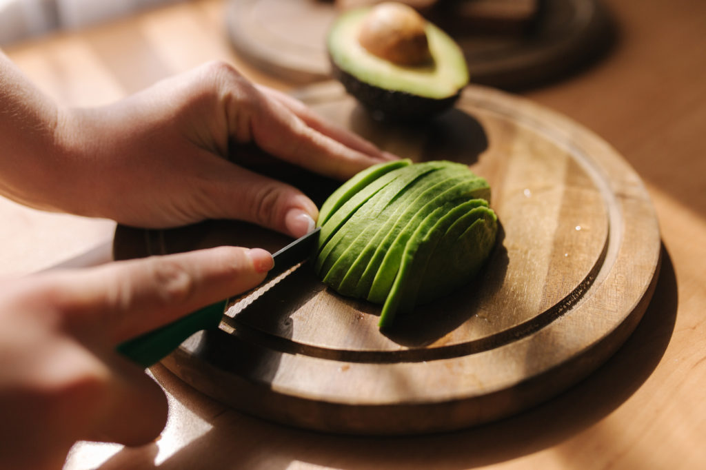 Close up of woman slicing avocado on wooden board at home. Vegetarian food concept.